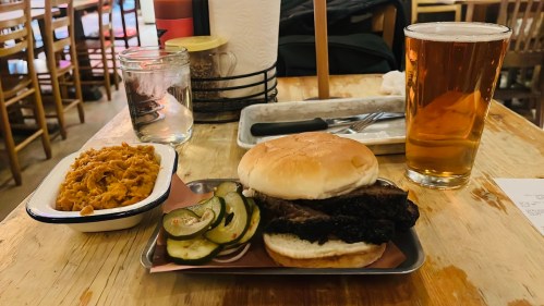 A plate with a sandwich topped with pickles, a side of macaroni, and a pint of beer on a wooden table.
