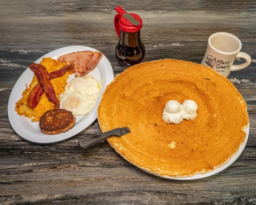 A large pancake with two scoops of ice cream, alongside a breakfast plate with eggs, bacon, sausage, and syrup.
