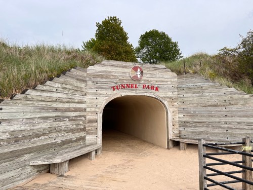 Entrance to Tunnel Park, featuring wooden steps and a rounded archway, surrounded by sandy dunes and greenery.