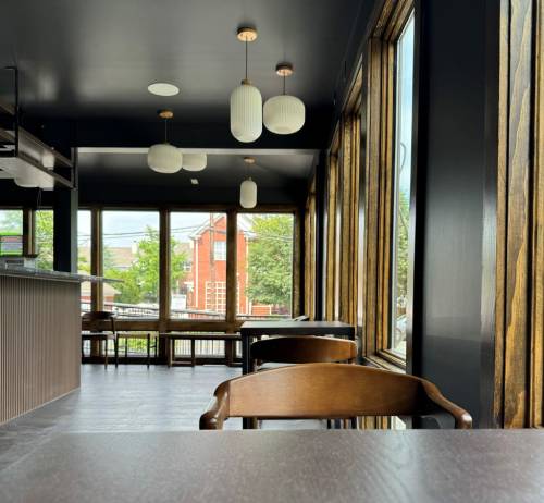 A modern café interior with wooden accents, large windows, and pendant lights, featuring empty tables and chairs.