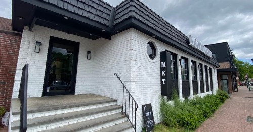 Exterior of a modern restaurant with a white brick facade, black accents, and a sign reading "MKT." Greenery in front.