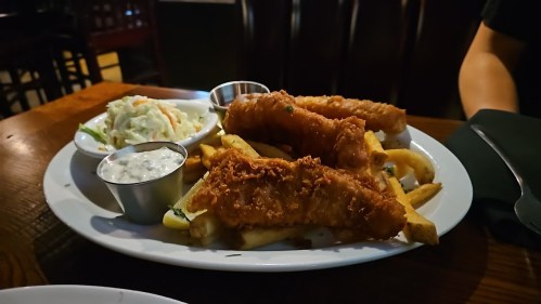 A plate of crispy fried fish fillets, French fries, coleslaw, and dipping sauce on a wooden table.