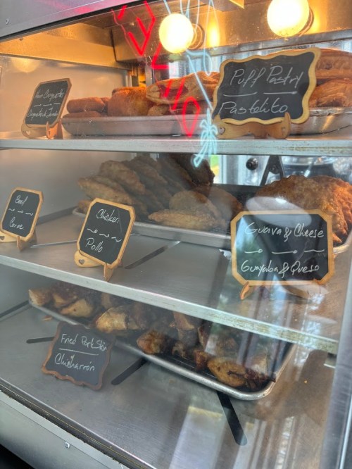 Display case filled with various pastries, including chicken, guava and cheese, and fried plantains, labeled on chalkboard signs.