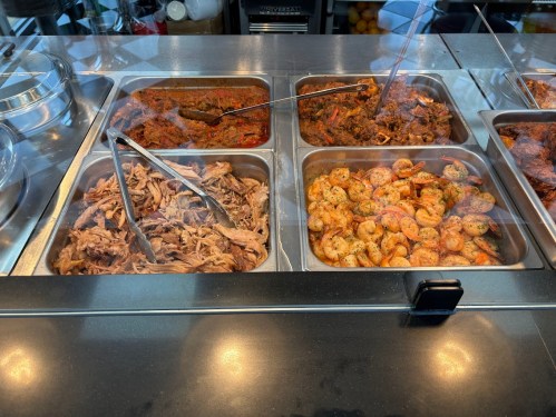 A display of various cooked dishes, including pulled meat and seasoned shrimp, in metal trays with serving utensils.