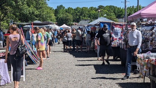 A bustling outdoor market with vendors and shoppers browsing colorful stalls under a clear blue sky.