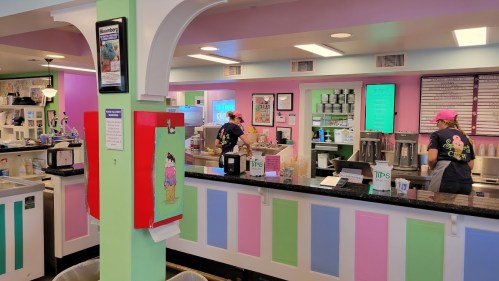 A colorful ice cream shop interior with staff serving customers at the counter and a tip jar visible.