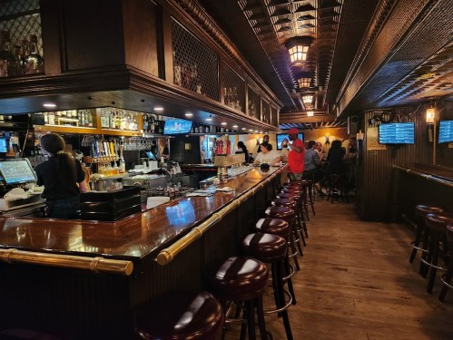 A cozy bar interior with wooden decor, a long counter, and patrons enjoying drinks in a warm, inviting atmosphere.