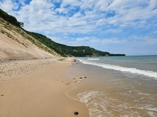 A serene beach scene with gentle waves, sandy shore, and lush green cliffs under a partly cloudy sky.
