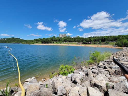A scenic view of a lake with a sandy beach, surrounded by greenery and a blue sky with fluffy clouds.