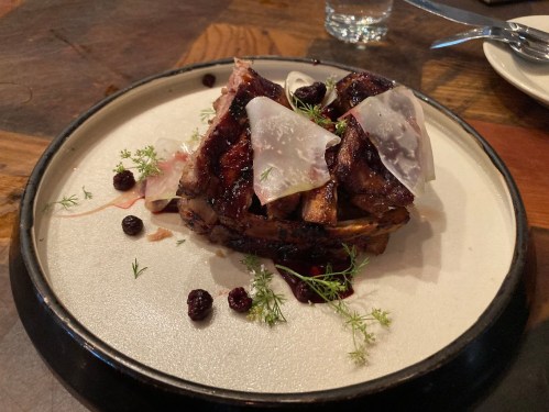 A plated dish featuring grilled meat with garnishes, topped with thin slices of cured meat and small berries.