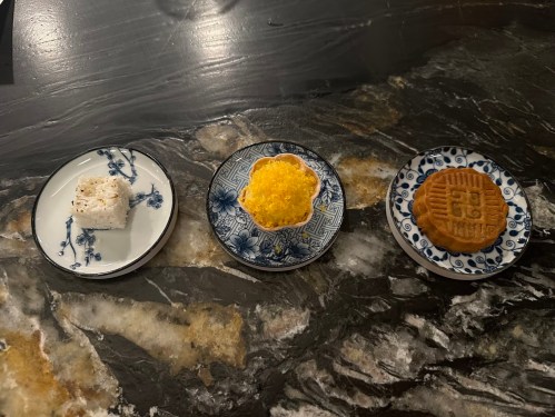 Three decorative plates on a marble surface, each with a different dessert: a white square, a yellow round, and a patterned cookie.