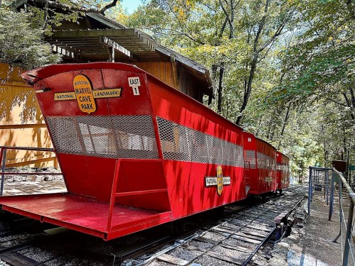 A red train car at Marvel Cave Park, surrounded by trees and a wooden structure in the background.