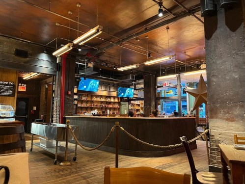 Interior of a bar with a wooden counter, shelves of bottles, and two screens displaying images. Warm lighting and rustic decor.