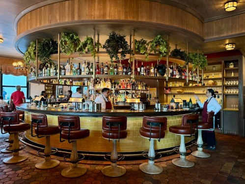 A stylish bar with a circular counter, shelves of liquor, and bartenders in aprons, surrounded by greenery and warm lighting.