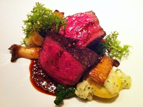 A beautifully plated dish featuring pink beef, mushrooms, greens, and a rich sauce on a white plate.