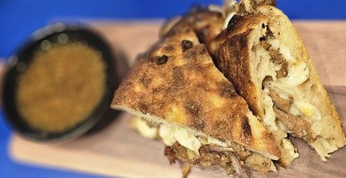 A close-up of a toasted sandwich filled with meat and melted cheese, served with a small bowl of sauce on the side.