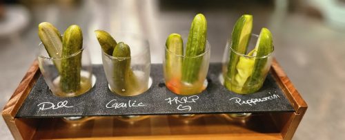 Four types of pickles in test tubes labeled Dill, Garlic, FRRP, and Peppercorn, displayed on a wooden stand.