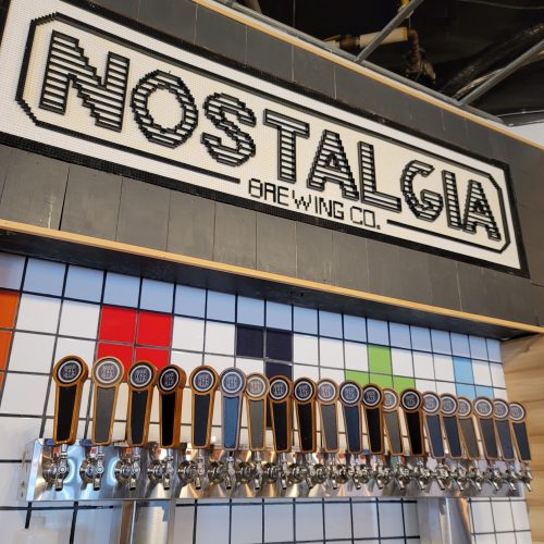 A colorful brewery tap wall with multiple beer taps and a sign reading "Nostalgia Brewing Co." above.