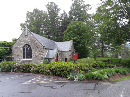 A stone church surrounded by greenery, with a red door and a sign, on a rainy day.