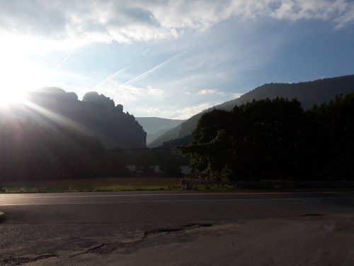 Sunrise over mountains, with a clear sky and soft clouds, casting light on a serene landscape and a winding road.