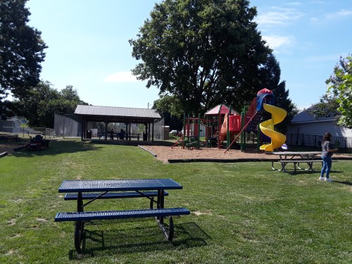 A playground with a slide and climbing structure, picnic tables, and a shelter in a grassy park on a sunny day.