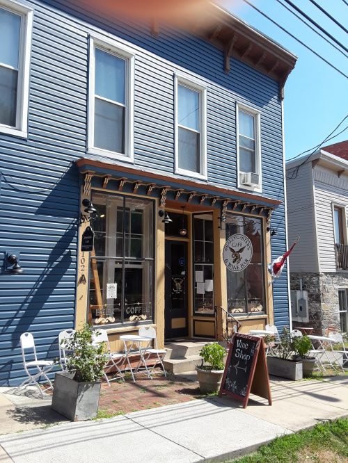A blue building with a café entrance, outdoor seating, and a sign for a wine shop. Flower pots line the walkway.