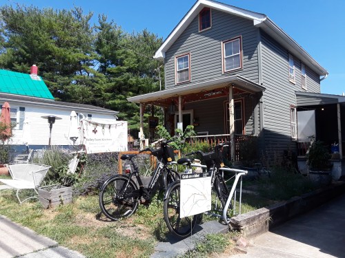 A gray house with a porch, surrounded by greenery, featuring two bicycles parked in front and a sign for a farm kitchen.