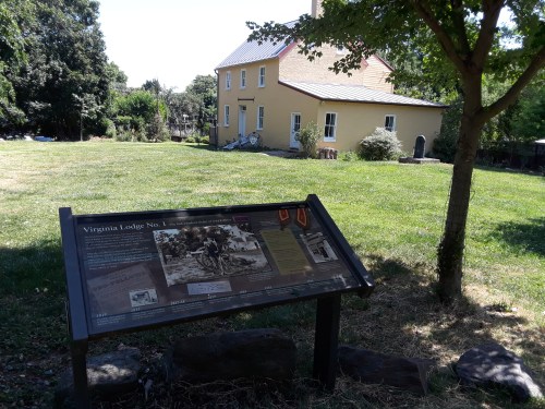 A yellow house with a grassy area and a historical sign in the foreground, surrounded by trees.