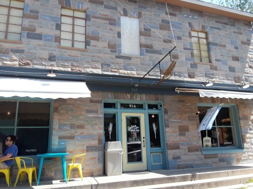 A stone building with a bakery entrance, featuring a sign, outdoor seating, and large windows.