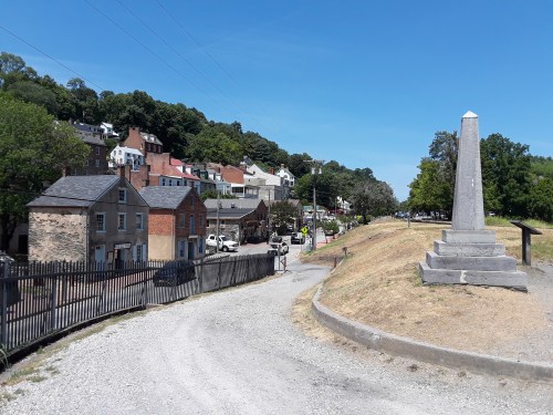 A gravel path leads to a monument, with historic buildings and trees in the background under a clear blue sky.