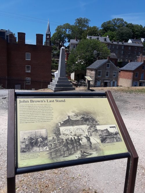 Sign about John Brown's Last Stand with an obelisk in the background and historic buildings under a clear blue sky.