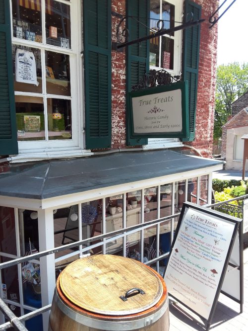 A quaint storefront with green shutters, a sign for "True Treats," and a wooden barrel outside.