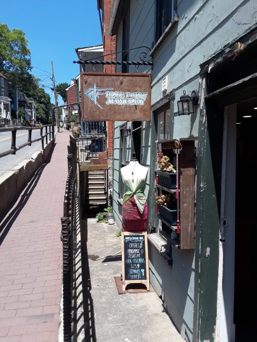 A storefront with a sign reading "Blue Point Jewelry" and a display mannequin outside, surrounded by greenery.