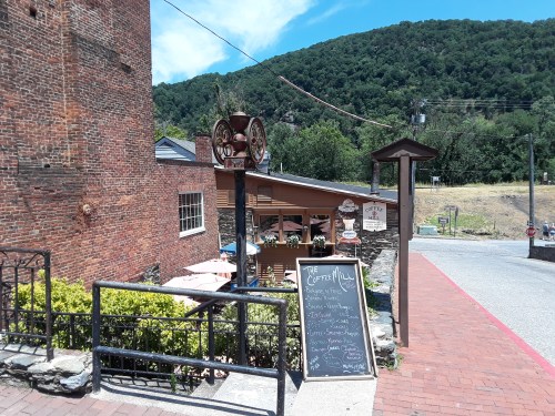 A quaint street scene featuring a café with outdoor seating, surrounded by greenery and a mountain backdrop.