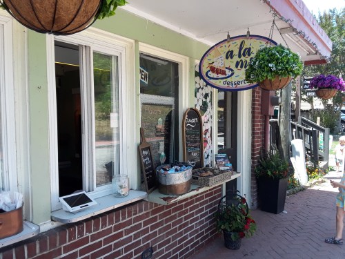 A quaint dessert shop with a sign, flower baskets, and a child standing outside on a sunny day.