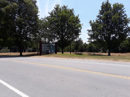 Sign for Harpers Ferry National Historical Park beside a road, framed by trees and open grassy areas under a clear sky.