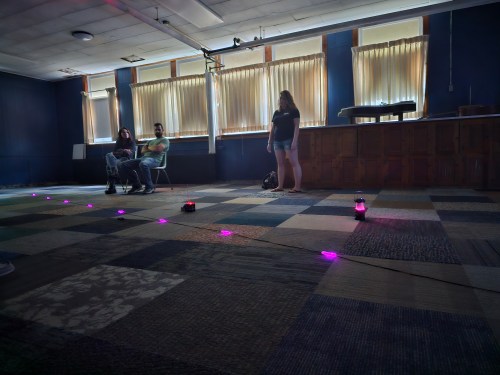 A dimly lit room with three people, two sitting on chairs and one standing, near glowing purple lights on the floor.