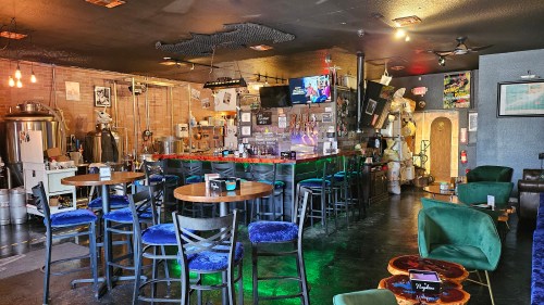 A cozy bar interior with wooden tables, high chairs, and a colorful counter, featuring eclectic decor and dim lighting.