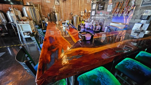 A wooden bar with a glossy finish, colorful stools, and brewing equipment in a brewery setting.