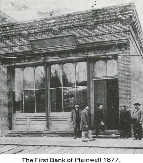 Historic photo of the First Bank of Plainwell, 1877, featuring a brick building and a group of men outside.