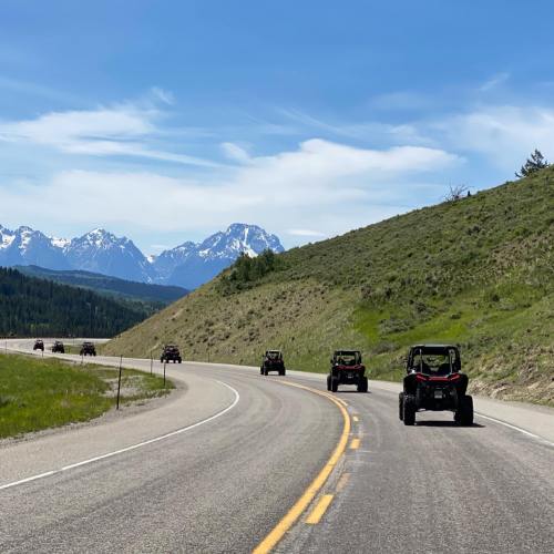 A line of off-road vehicles driving along a winding road with mountains in the background under a clear blue sky.