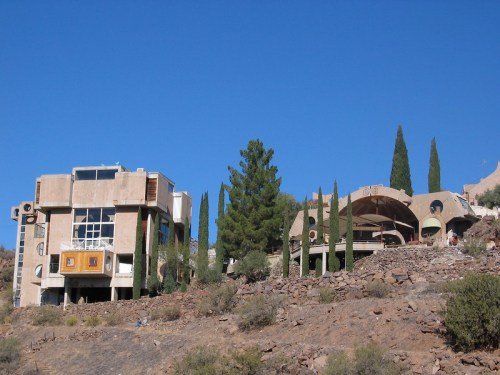 Unique architectural structures on a hillside, surrounded by tall cypress trees and clear blue sky.