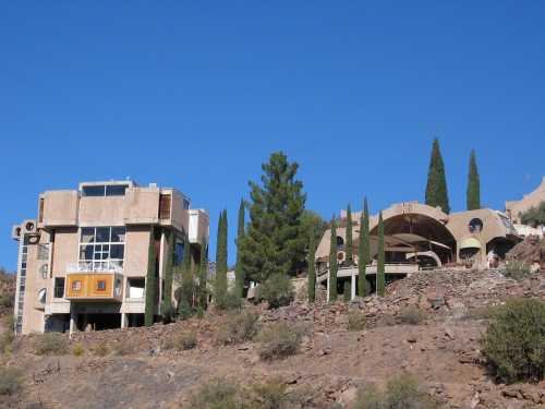 Unique architectural structures on a hillside, surrounded by tall cypress trees and clear blue sky.