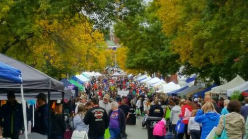 A bustling street fair with colorful tents and a large crowd under trees with autumn leaves.