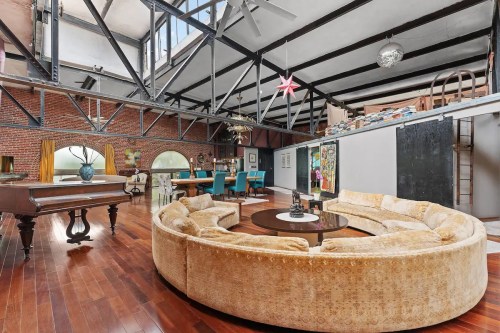 Spacious loft with high ceilings, exposed beams, circular sofa, and a grand piano, featuring a mix of modern and vintage decor.