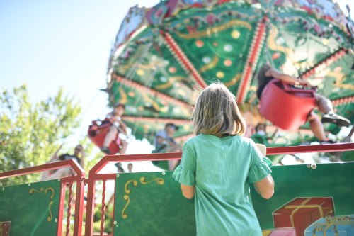 A child watches a colorful swing ride with excited riders above, surrounded by greenery on a sunny day.