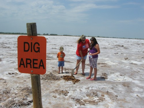 A family digs in a salt flat, with a "Dig Area" sign nearby and a clear sky in the background.