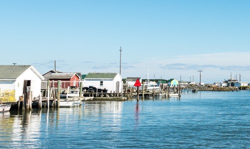 Colorful houses line a calm waterfront, reflecting in the water under a clear blue sky.