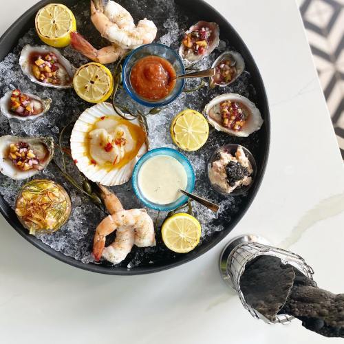 A seafood platter with shrimp, oysters, scallops, sauces, and lemon slices on ice, served in a round black dish.