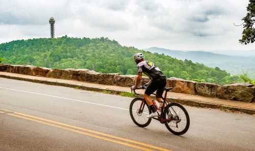 A cyclist rides along a scenic road with a tower in the distance, surrounded by lush green hills under a cloudy sky.